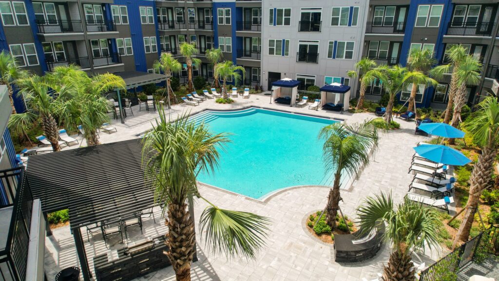 Aerial view of an apartment complex courtyard featuring a central pool with surrounding lounge chairs, cabanas, palm trees, and a pergola-covered sitting area.