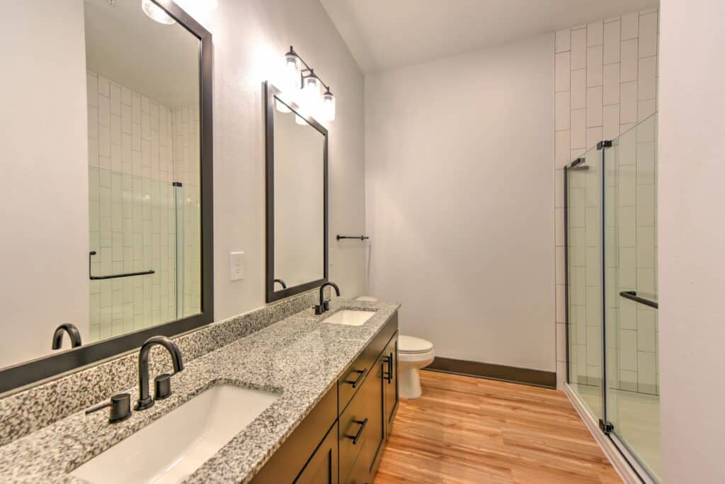 Bathroom with a double vanity, granite countertops, large mirrors, and a glass-enclosed shower. The floor is wood-style, and there's a toilet next to the shower.