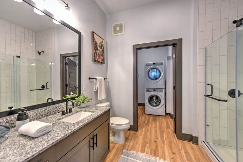 Modern bathroom with granite countertop, single sink, and large mirror. A stacked washer and dryer are visible in an adjacent laundry area. Glass shower door and wood flooring.