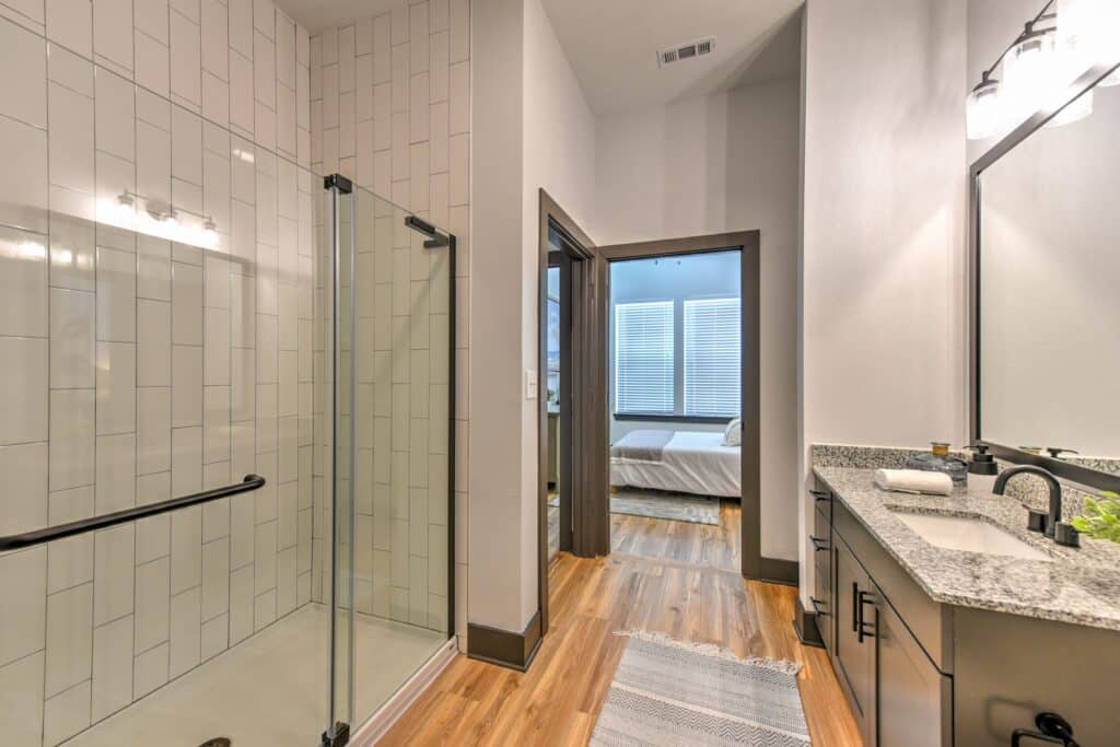 Modern bathroom with a glass shower, granite countertop, single sink, and wooden floor. A view into a bedroom with a bed is visible through an open door.