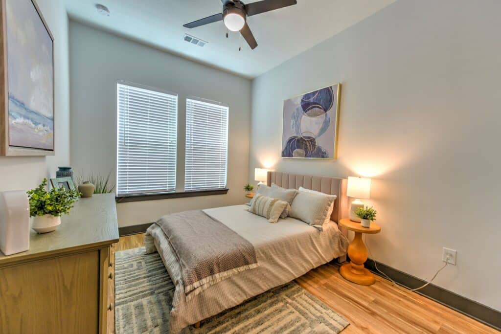 A cozy bedroom features a bed with light-colored bedding, two side tables with lamps, a dresser with plants, a ceiling fan, and a large window with blinds. Artwork hangs above the bed.