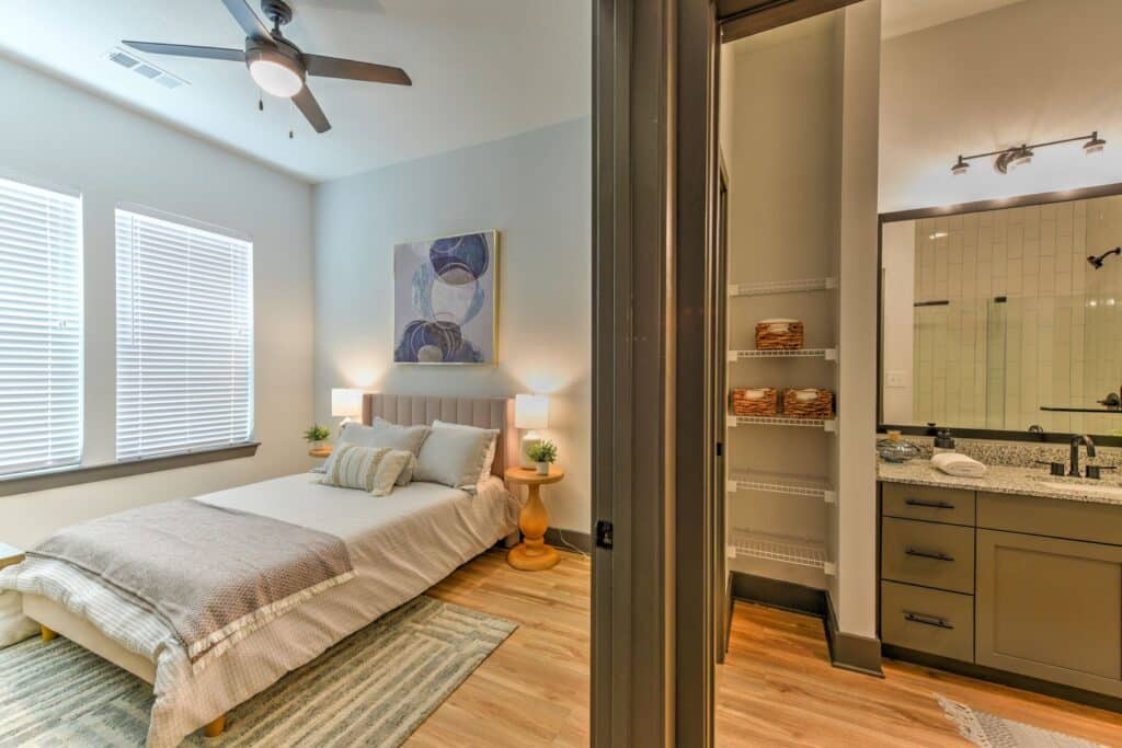 A modern bedroom with a bed, pillows, and art on the wall, adjacent to a bathroom with a granite countertop and shelves stocked with towels.