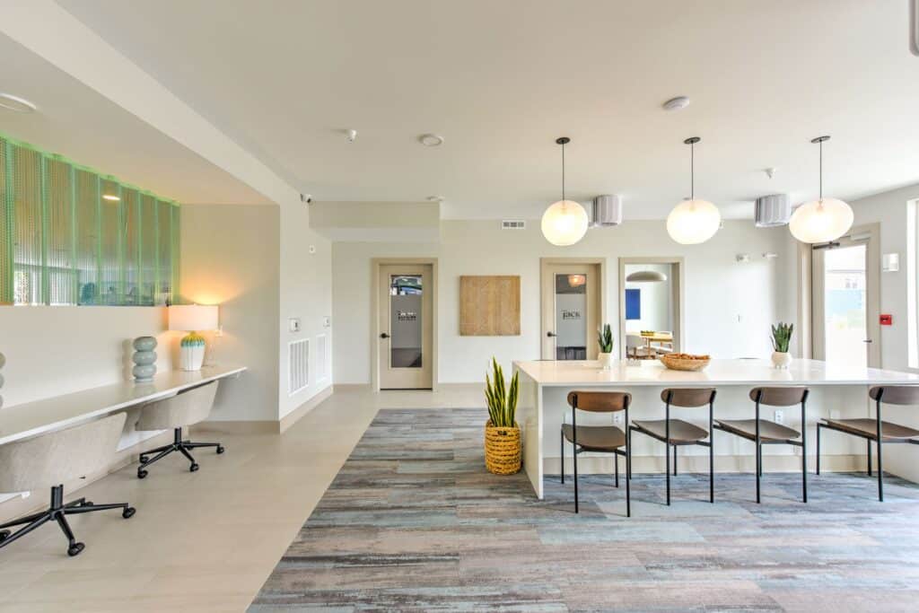 Modern communal workspace with a long table, four chairs, pendant lights, a decorative plant, and desks with chairs on the left.