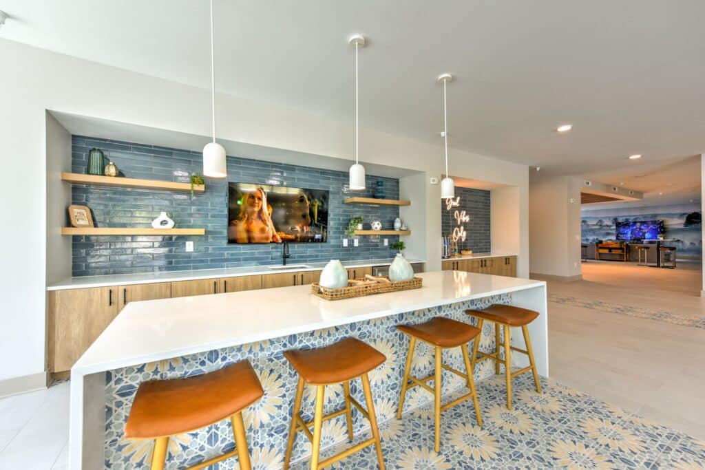 Modern kitchen with a white island, wooden stools, decorative tile backsplash, and a wall-mounted TV. Pendant lights hang above the island.