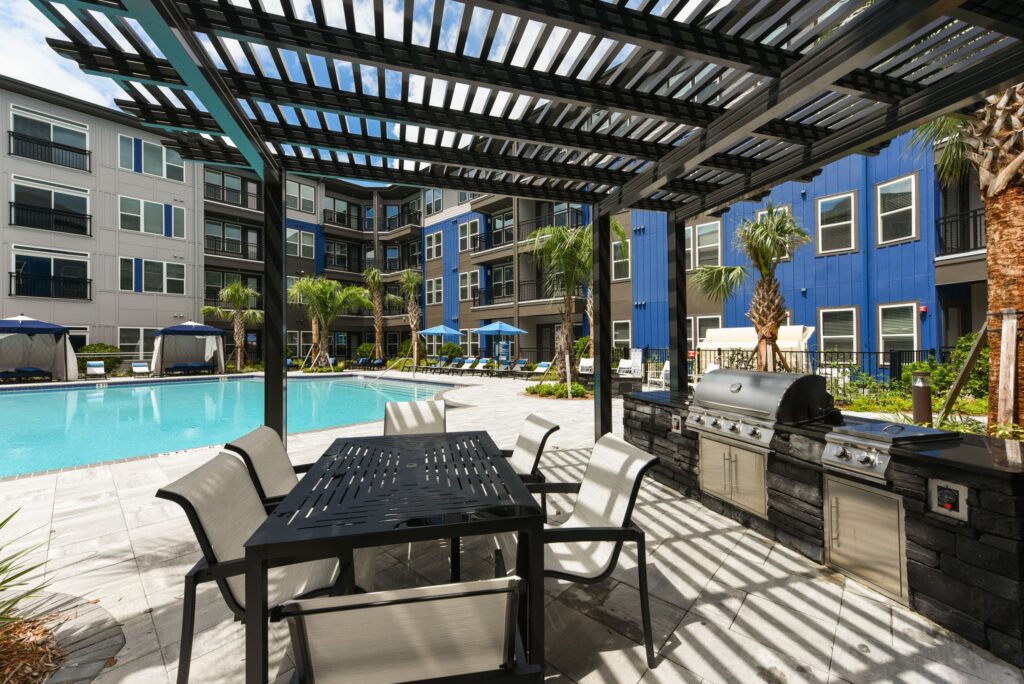 Outdoor pool area with a shaded dining table and grill, surrounded by modern apartment buildings and palm trees.