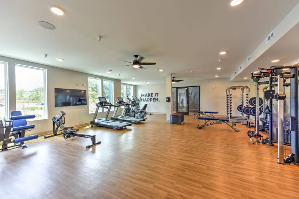 A spacious gym with exercise equipment, including treadmills, weight machines, and a stationary bike. Large windows and a wall with the words "MAKE IT HAPPEN.