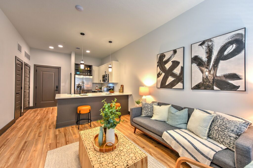 Modern living room with a gray sofa, patterned cushions, a wooden coffee table with plants, abstract art on the wall, and a view into a kitchen with bar seating and stainless steel appliances.