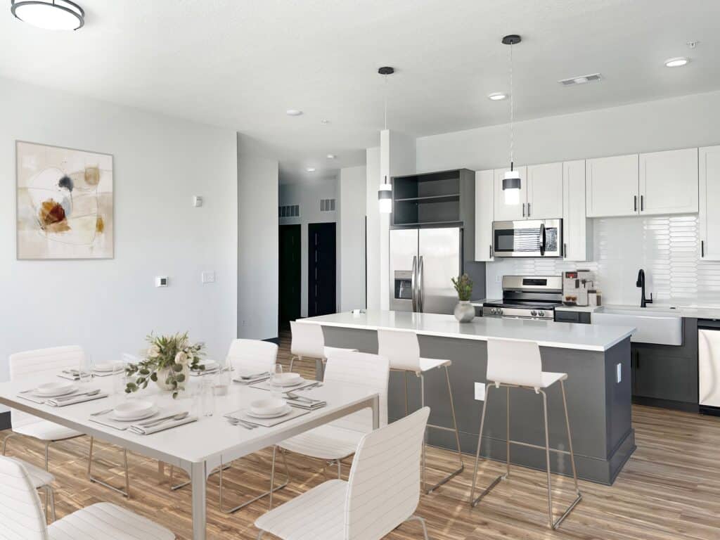 Modern kitchen and dining area with a white table, white chairs, and a black and white kitchen island. Stainless steel appliances and contemporary light fixtures are visible.