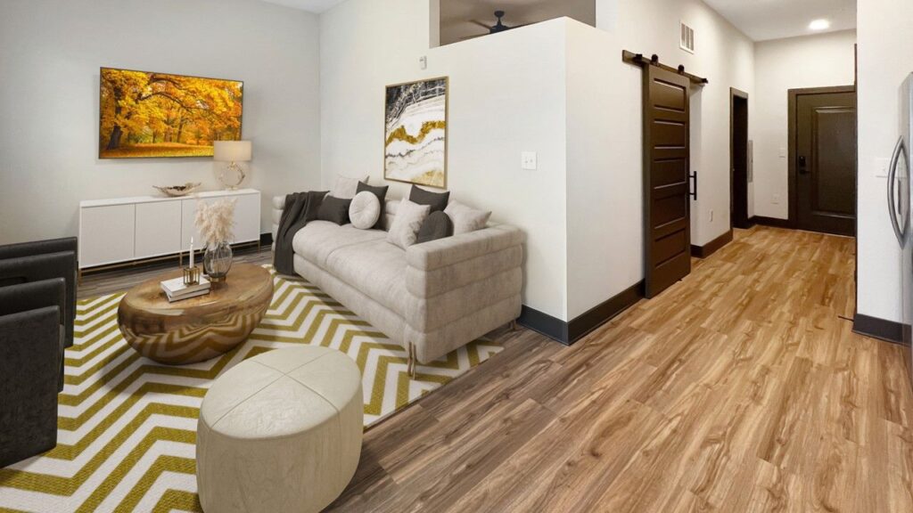 Living room with a beige sofa, patterned rug, round wooden table, and wall artwork. A sliding door leads to a hallway with wood flooring. A lamp sits on a sideboard.