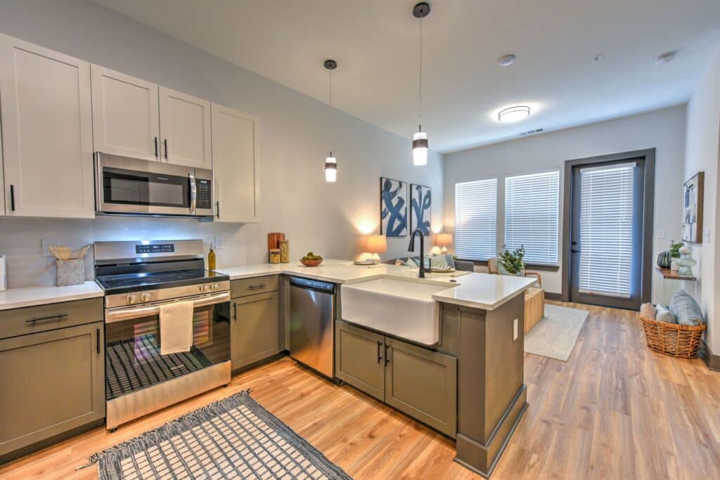 Modern kitchen with stainless steel appliances, white and gray cabinetry, farmhouse sink, and hardwood floors. Pendant lights hang above the island, and large windows provide natural light.