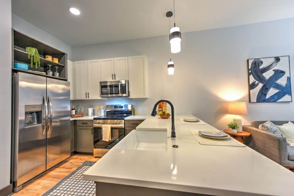 Modern kitchen with stainless steel appliances, white cabinets, and a long island countertop. Pendant lights hang above, and there's a blue abstract painting on the wall.