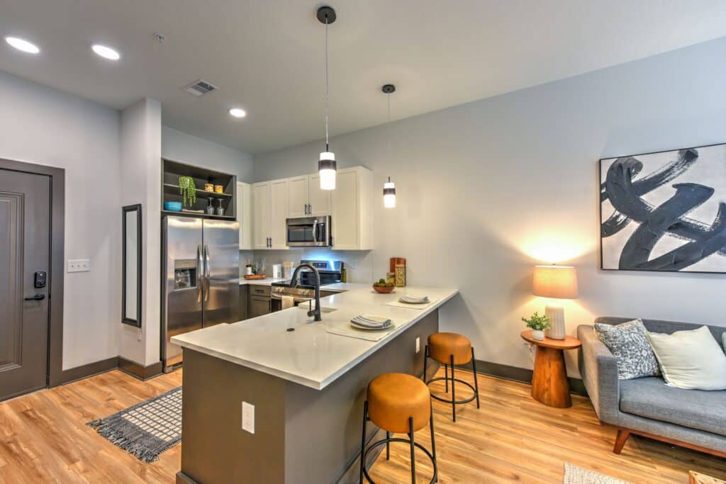 Modern kitchen and living area with a central island, pendant lights, stainless steel appliances, a gray sofa, and abstract wall art.