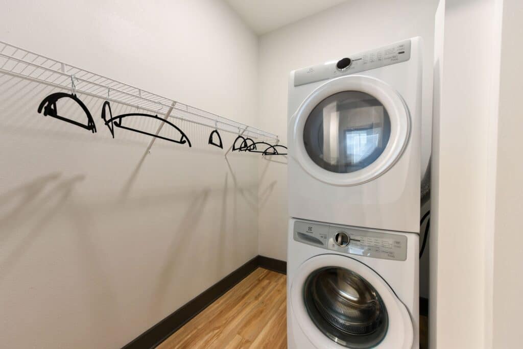 Laundry room with a stacked washer and dryer on the right, and an empty wire shelf with hangers on the left. The floor is wooden and the walls are light-colored.