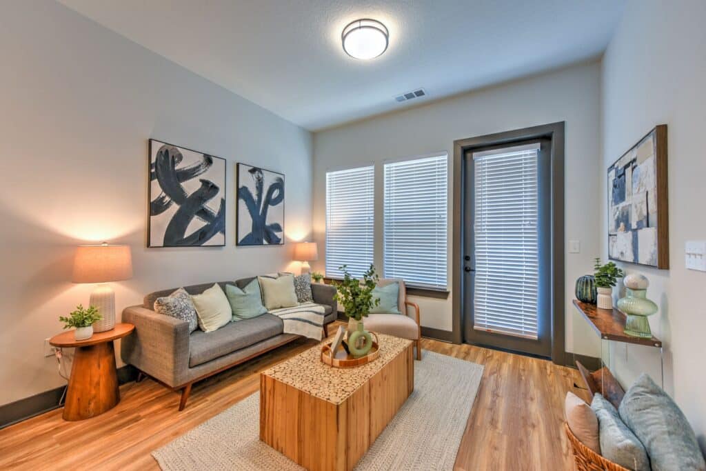 A cozy living room with a gray sofa, wooden coffee table, and two abstract paintings on the wall. Two lamps illuminate the space, and a large window with blinds adds natural light.