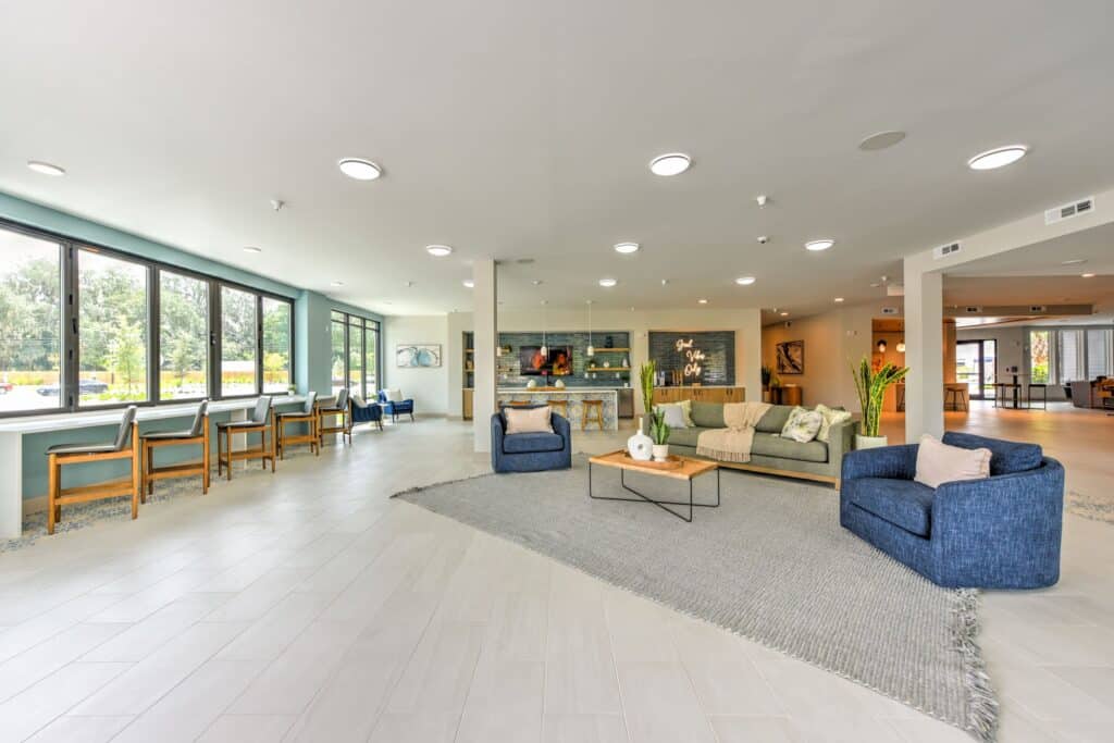 Spacious, modern lounge area with large windows, two blue armchairs, a sofa, and a coffee table on a gray rug. Tables and chairs line the far wall.