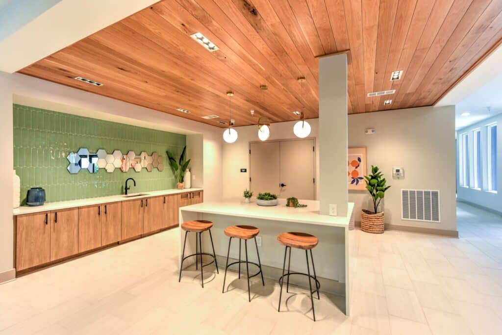 Modern kitchen with wooden ceiling and cabinetry, green backsplash, and three wooden stools at a white island. Three circular pendant lights hang overhead, and plants add a touch of greenery.