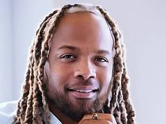 Man with dreadlocks smiling, wearing a light shirt, close-up view.