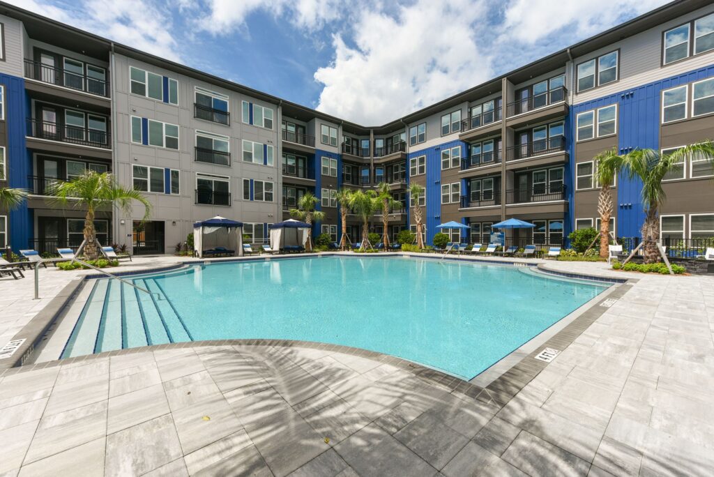 Modern apartment complex with a central pool, surrounded by lounge chairs, umbrellas, and palm trees, under a partly cloudy sky.