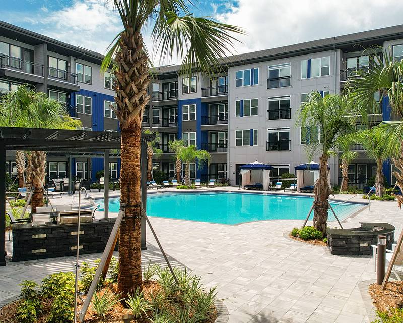 A modern apartment complex with a central swimming pool, surrounded by palm trees and sun loungers, under a partly cloudy sky.