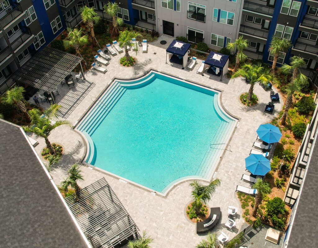 Aerial view of a square swimming pool surrounded by lounge chairs, umbrellas, and palm trees in an apartment complex courtyard.