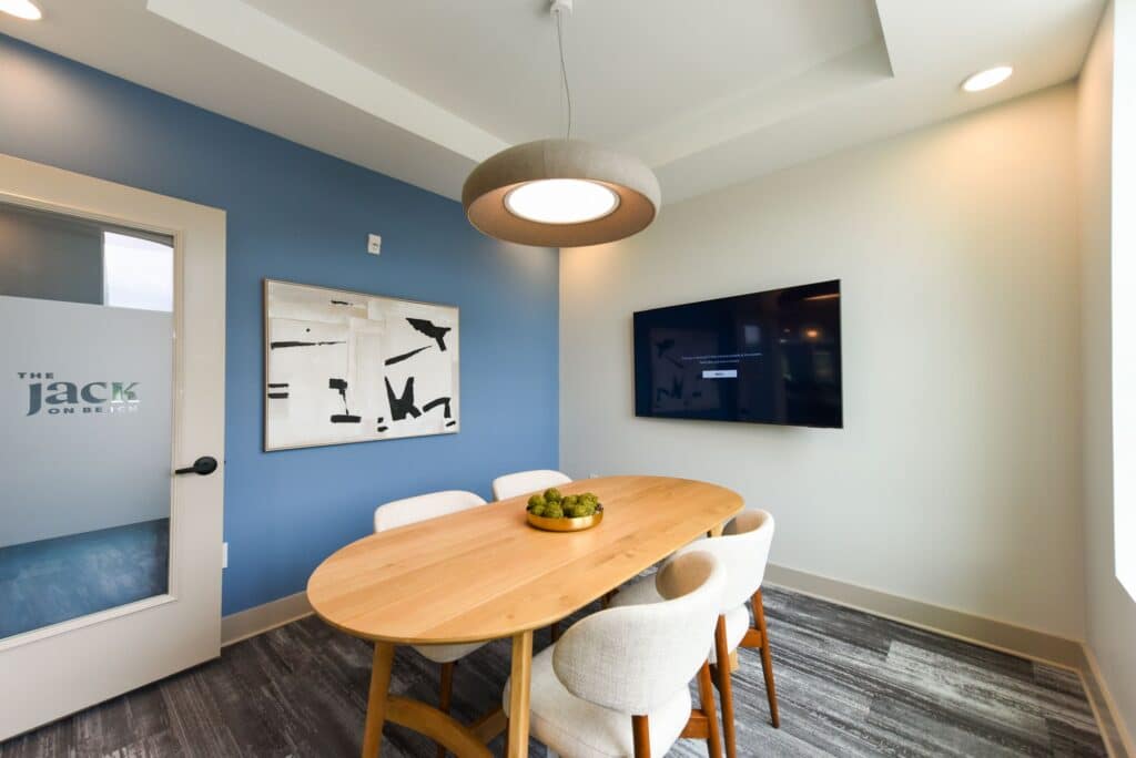 A small conference room with a wooden table, four chairs, a wall-mounted TV, abstract wall art, a bowl of green apples on the table, and a round ceiling light.