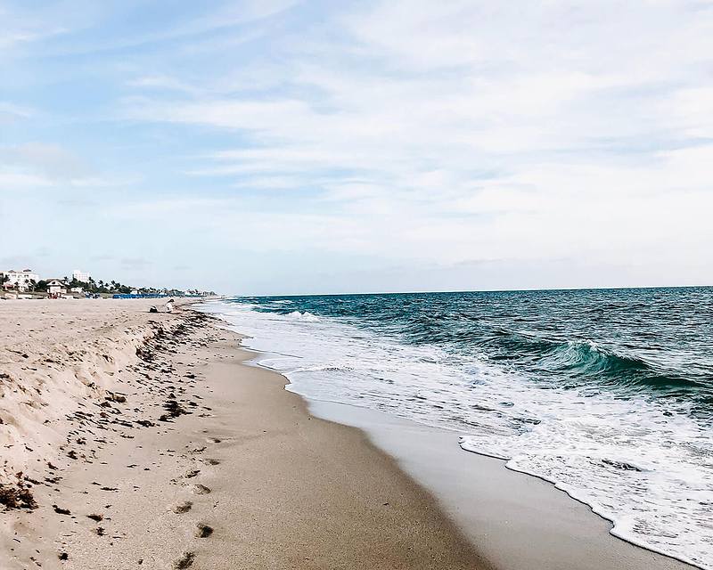 A sandy beach with gentle waves and a clear blue sky, featuring footprints in the sand and a distant line of buildings.